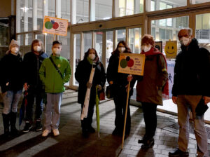 Gruppe mit Maske auf dem Protest gegen von Querdenkern organisierte Montagsdemos. Im Hingergrund der hell erleuchte Bahnhof, davor die Gruppe Gegendemonstranten, darunter Sabine Gruetzmacher mit Maske und Protestschildern.