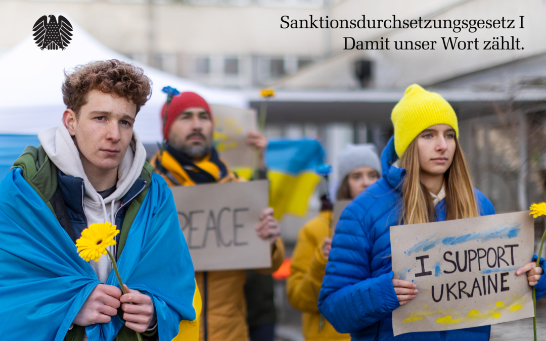Eine Gruppe Protestierender Jugendlicher mit Schildern "Stop War" und "I stand for Ukraine" und gehüllt in die Ukrainische, blau-gelbe Flagge. Auf dem oberen Bildrand inks der Bundesadler, rechts die Worte "Sanktionsdurchsetzungsgesetz 1" und "Damit unser Wort zählt".
