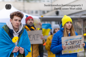 Eine Gruppe Protestierender Jugendlicher mit Schildern "Stop War" und "I stand for Ukraine" und gehüllt in die Ukrainische, blau-gelbe Flagge. Auf dem oberen Bildrand inks der Bundesadler, rechts die Worte "Sanktionsdurchsetzungsgesetz 1" und "Damit unser Wort zählt".