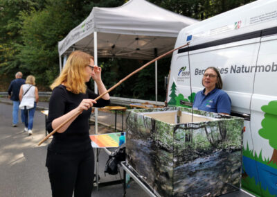 Sabine (links) in der Hand eine Angelrute, am Stand der Biologischen Station und des Bergischen Naturmobils. Eine Mitarbeiterin, dazwischen ein Kasten mit Flußtierchen zur Bestimmung der Wassergüte. Sabine Angelt einen "Strudelwurm".