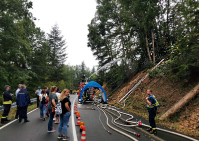 Eine Straße im Wald, eine bunte Gruppe BesucherInnen der Blaulichtmeile-Veranstaltung bestaunt einen Feuerwehr-Spritzenwagen, der in hohem Bogen Wasser an den rechten Hang in die Bäume schiesst