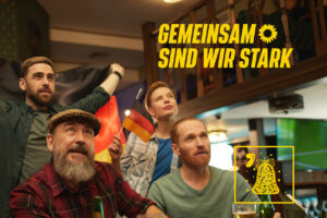 Eine Vierergruppe Menschen in einer Kneipenszene, ein Mann hinten hält eine Deutschlandfahne hoch.