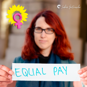 Sabine leicht verschwommen hält ein weißes Schild in die Kamera. Auf ihm steht in mintgrüner Farbe: Equal Pay. Links oben im Bild die typische gelbe Sonnenblume. In ihr ein pink Frauensymbol.