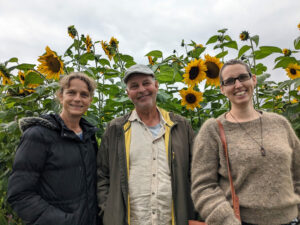 Vor einem Feld Sonnenblumen, stehen Anne Monika Spallek links, rechts Sabine und in der Mitte Seb Schäfer. Sie lächel in die Kamera während in ihrem Rücken wunderschöne Sonnenblumen blühen - so hoch, dass sie in etwa auf ihrer Kopfhöhe sind