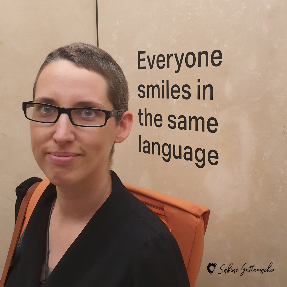 Sabine lächelt. Auf der beige Wand hinter ihr steht geschrieben 'everyone smiles in the same language' (jeder lächelt in derselben Sprache)